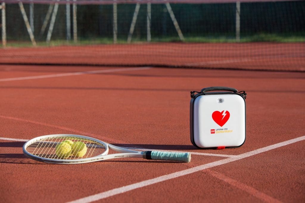 Lifepak CR2 Defibrillator on a tennis court next to a ball and racquet